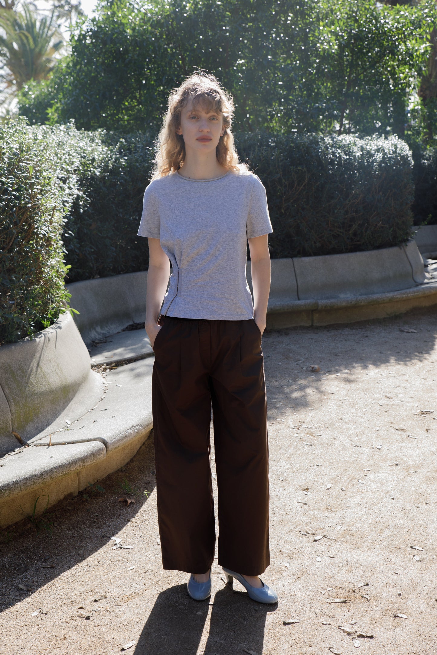 A woman with curly blonde hair stands outdoors on a sunny day, wearing a gray t-shirt and the Contrast Waist Pant - Brown by Deiji Studios. She is on a gravel path, surrounded by green bushes and trees.