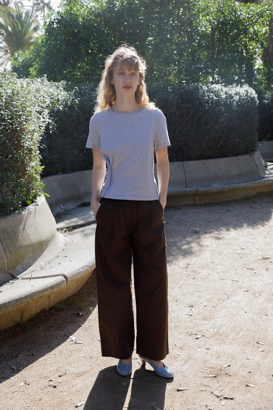 A woman with curly blonde hair stands outdoors on a sunny day, wearing a gray t-shirt and the Contrast Waist Pant - Brown by Deiji Studios. She is on a gravel path, surrounded by green bushes and trees.