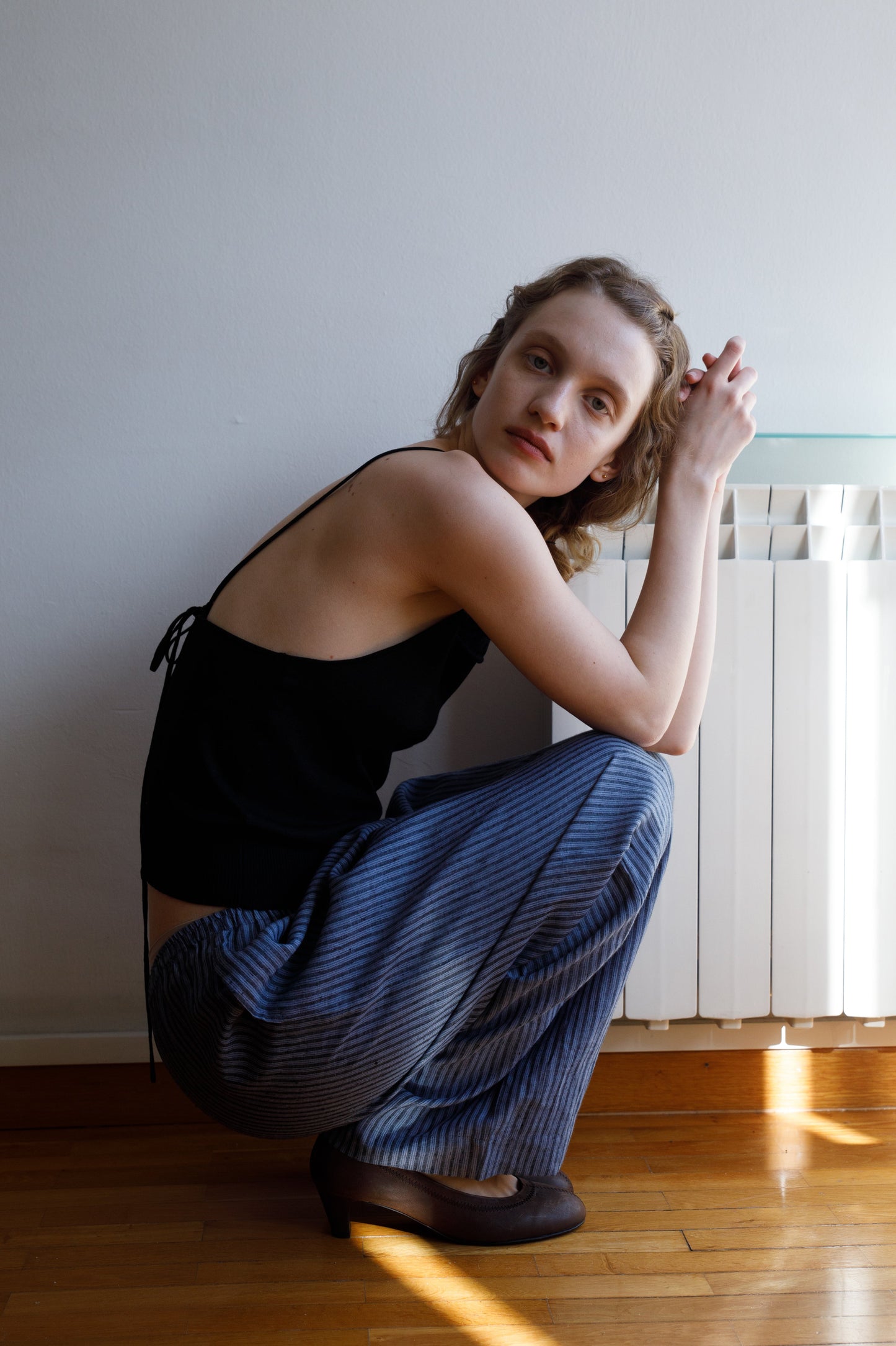 A woman with wavy light brown hair wears the Drawcord Knit Top - Black by Deiji Studios and blue striped pants as she squats on a wooden floor, leaning forward with her hands clasped, gazing softly at the camera.