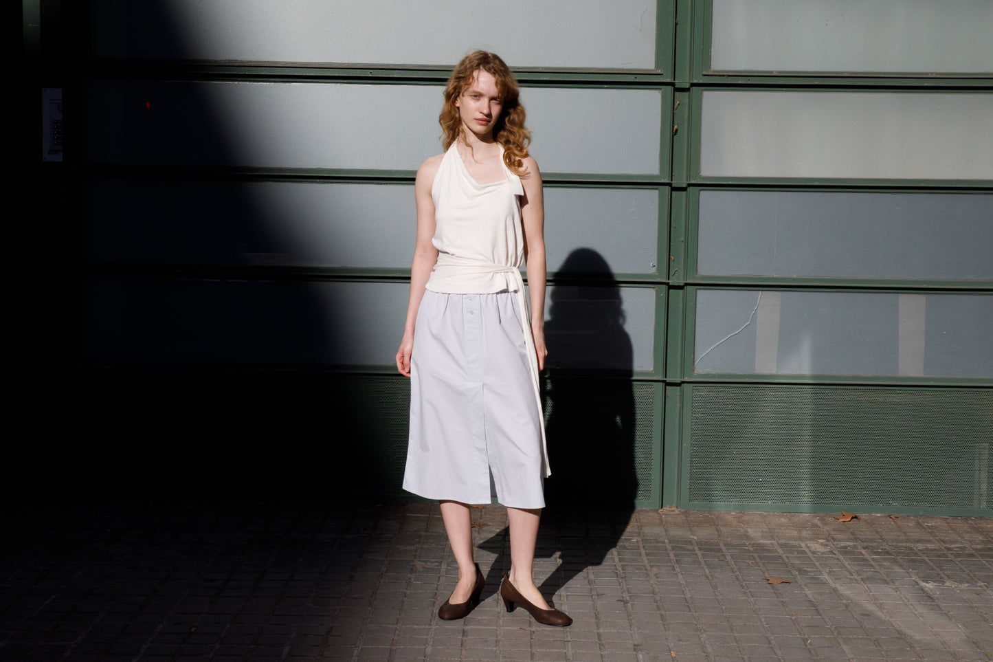 A woman with curly hair stands on a sidewalk by a green and frosted glass wall, wearing a sleeveless white top, the Overlap Skirt - Blue from Deiji Studios, and dark flats as sunlight and shadows fall across the scene.