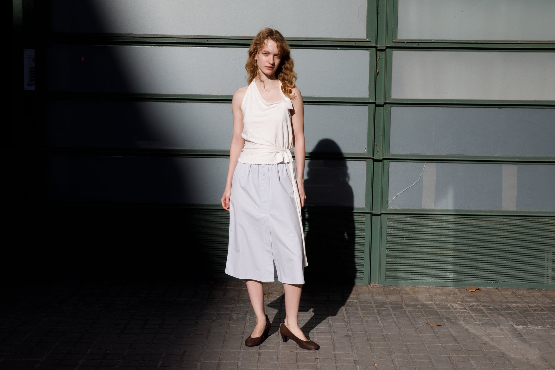 A woman with curly hair stands on a sidewalk by a green and frosted glass wall, wearing a sleeveless white top, the Overlap Skirt - Blue from Deiji Studios, and dark flats as sunlight and shadows fall across the scene.