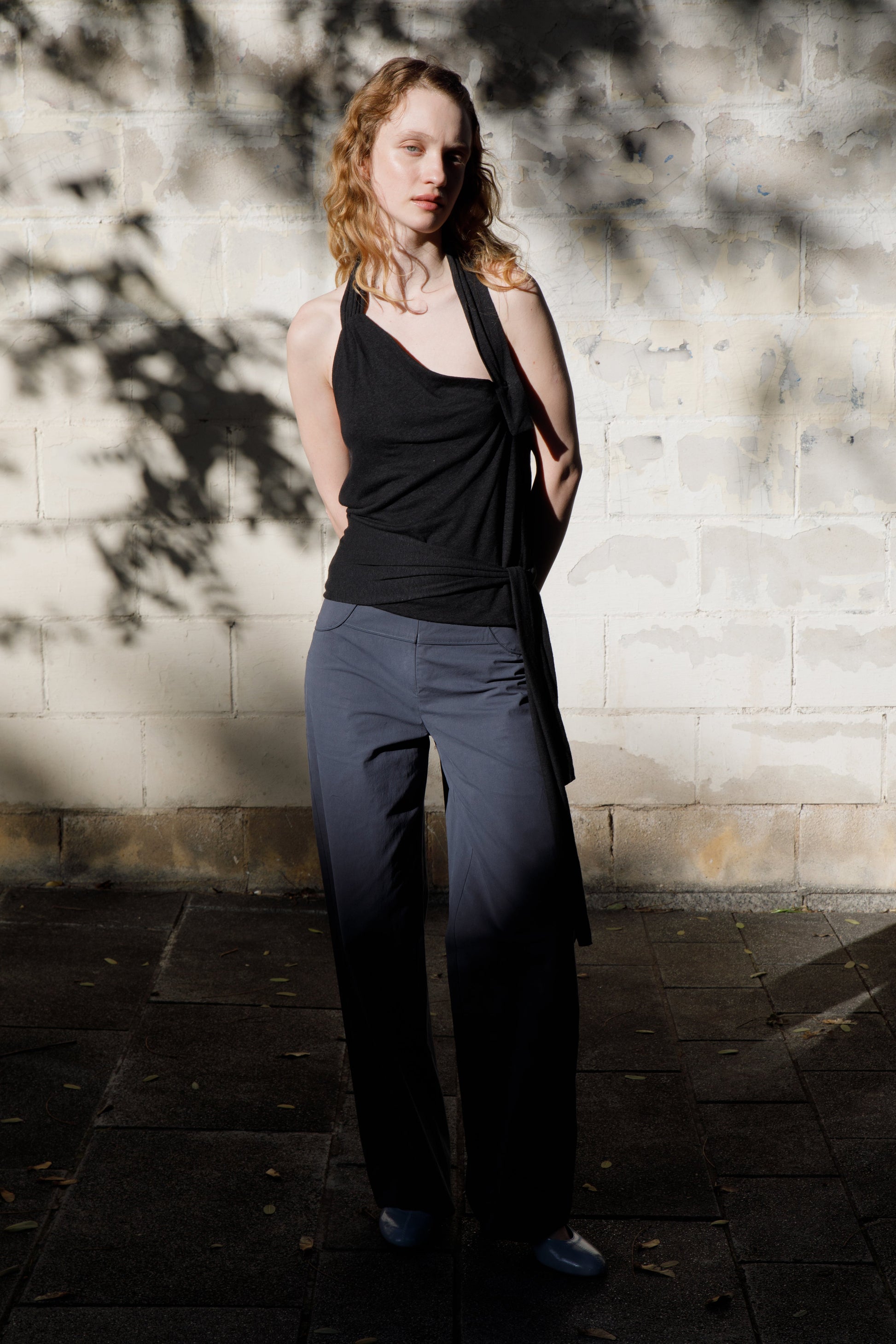 A person with wavy light hair stands against a sunlit brick wall, wearing a sleeveless black top and straight-leg Panelled Pant - Charcoal by Deiji Studios. Branch shadows fall across the scene.