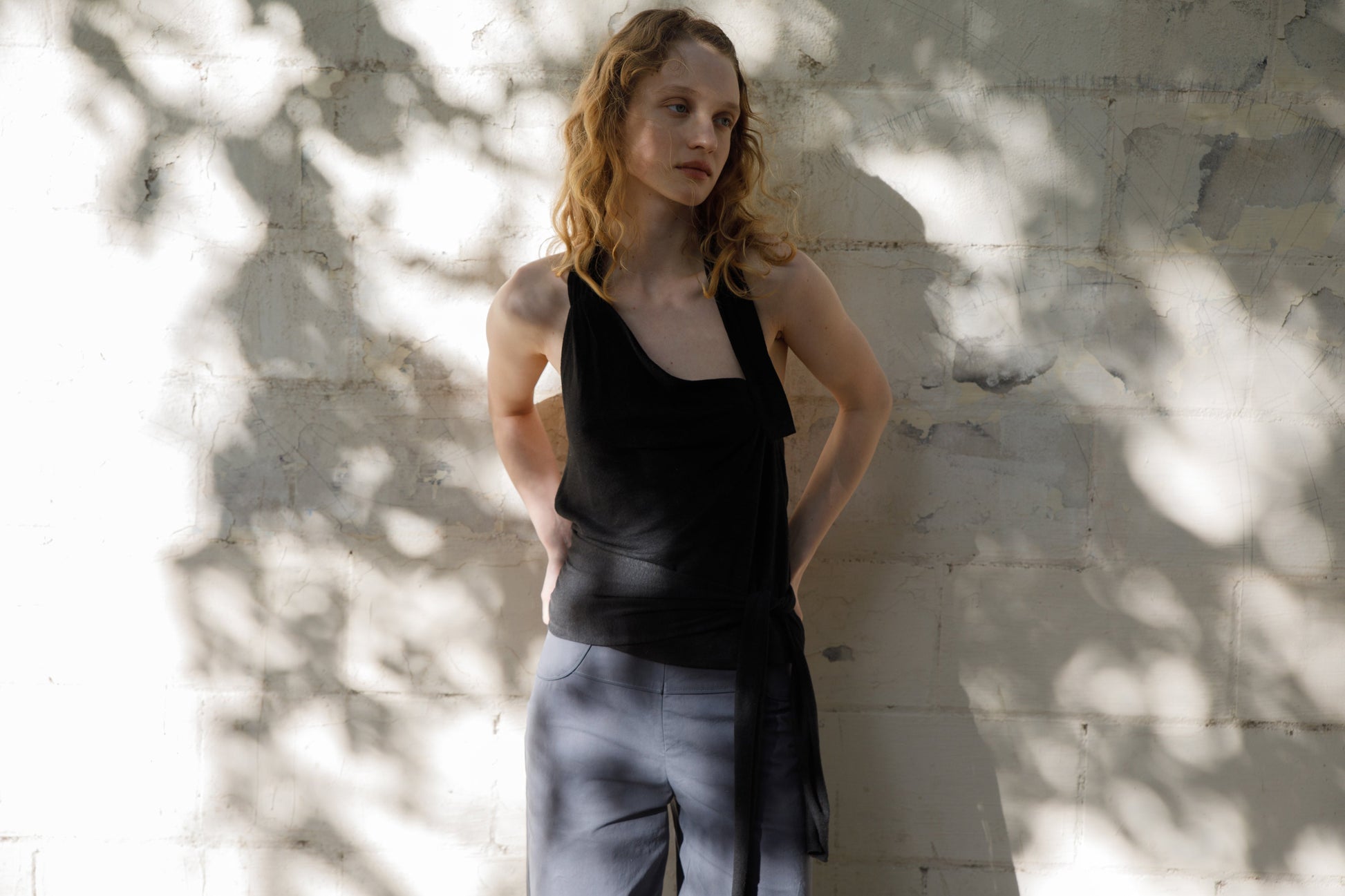 A woman with curly blonde hair stands against a sunlit, textured wall, wearing a sleeveless black top and the Panelled Pant - Charcoal by Deiji Studios. Shadows of tree leaves fall across both her and the wall.