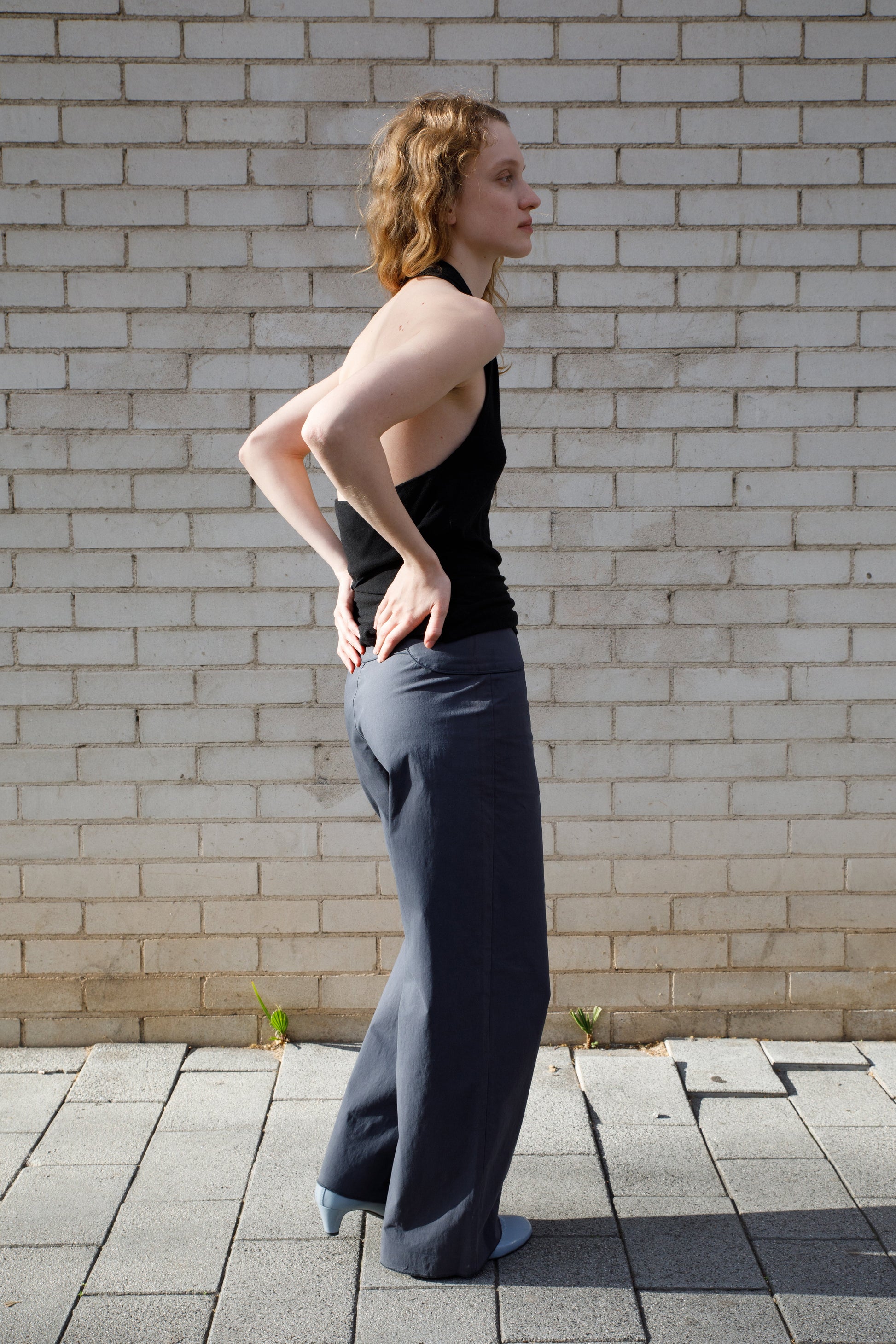A person with wavy light brown hair stands sideways on a paved walkway in front of a white brick wall, wearing a black sleeveless top, Deiji Studios Panelled Pant - Charcoal with a straight-leg silhouette, and light blue shoes.