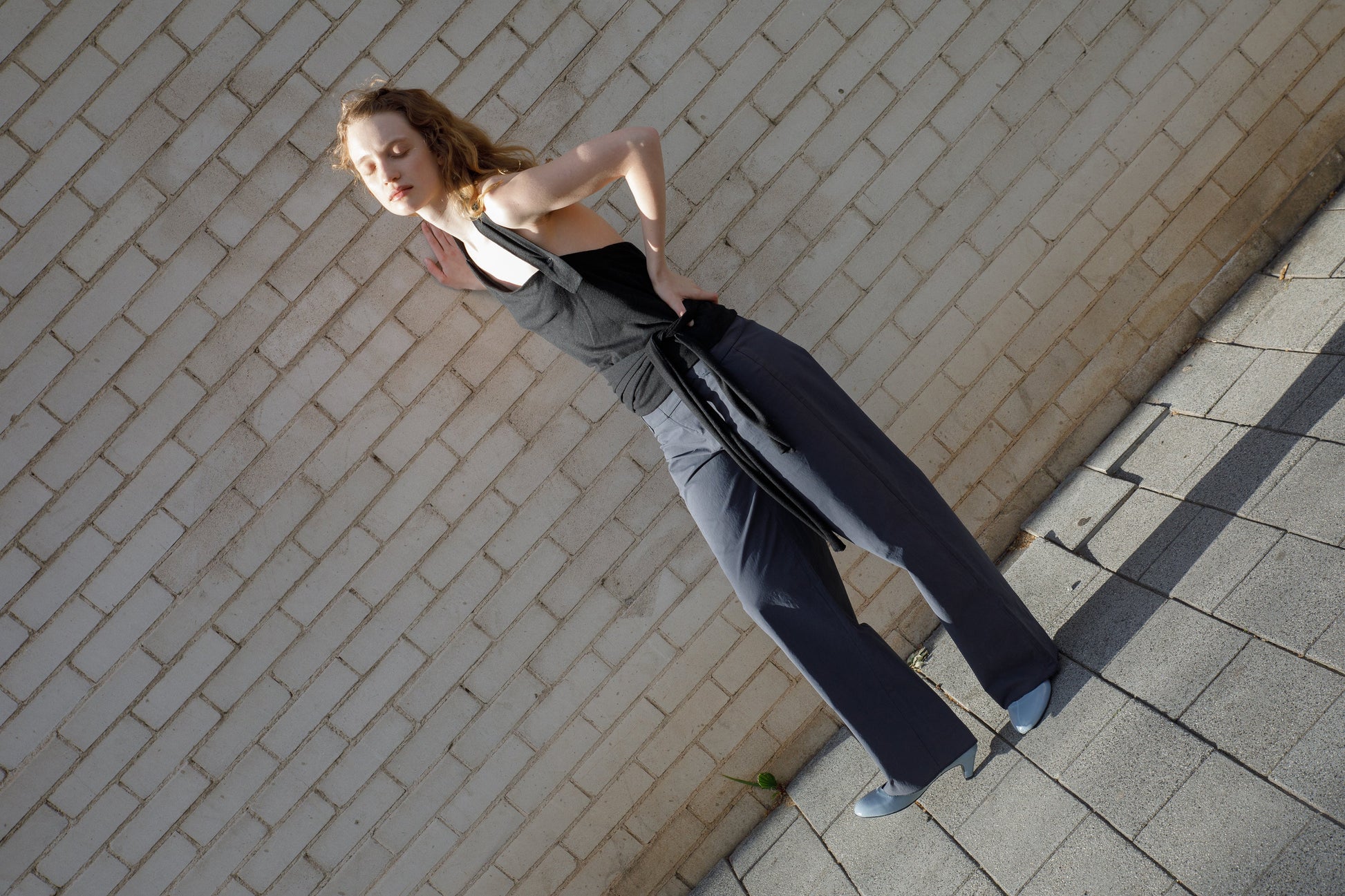 A person with wavy hair, in a black sleeveless top and Deiji Studios Panelled Pant - Charcoal, blue shoes, leans against a white brick wall on a sunlit sidewalk, eyes closed.