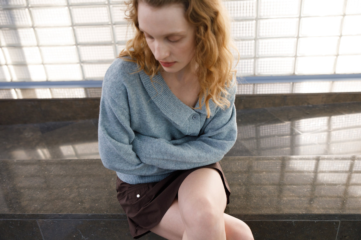 A woman with wavy, light brown hair sits on a stone bench, looking down with her arms crossed. She wears a light blue sweater and the Pocket Skirt - Sepia from Deiji Studios, with a glass block wall in the background.