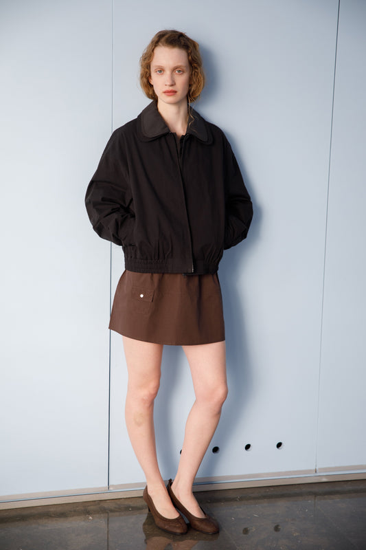 A person with short, wavy hair stands against a light blue wall, wearing a Deiji Studios Collared Zip Jacket - Black Contrast, a brown mini skirt, and brown flats, hands in jacket pockets and a neutral expression.