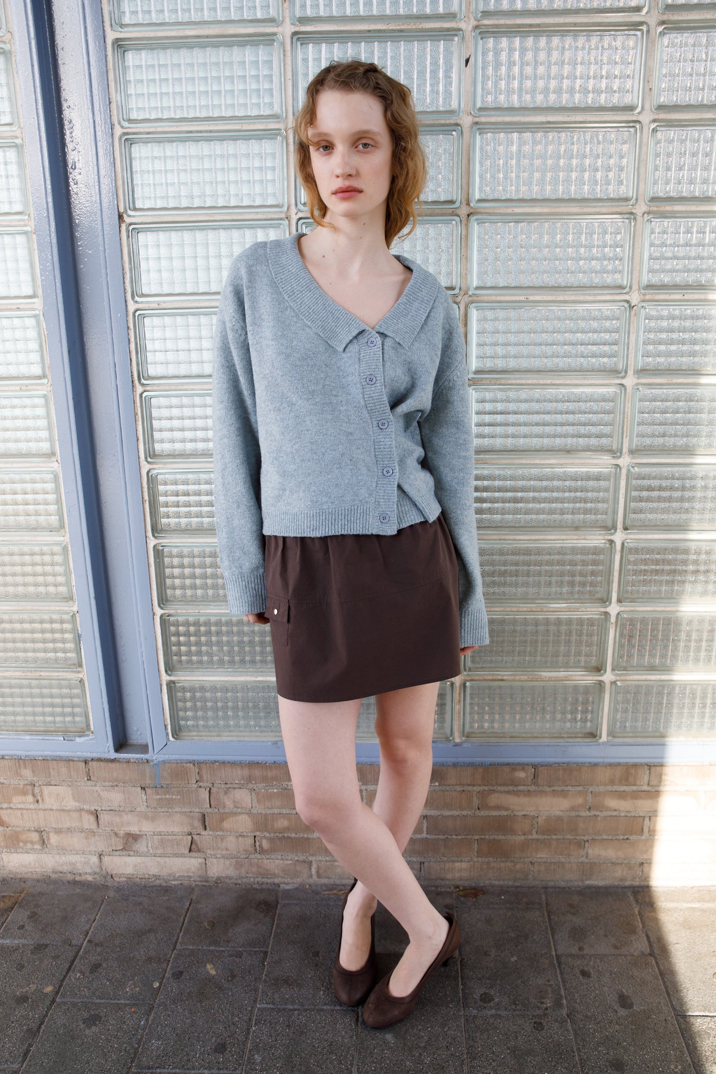 A young woman with wavy hair and brown flats stands before a glass brick wall, wearing a light blue cardigan and the Pocket Skirt - Sepia by Deiji Studios. She crosses her ankles and gazes neutrally ahead.