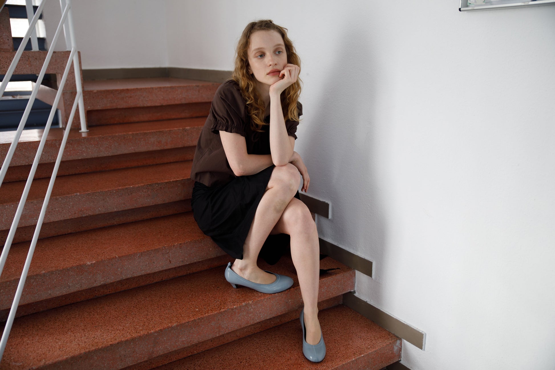 A woman with long, wavy hair sits on indoor stairs, resting her chin on her hand and gazing thoughtfully into the distance. She wears the Close Shirt in Sepia by Deiji Studios, a black skirt, and light blue shoes.