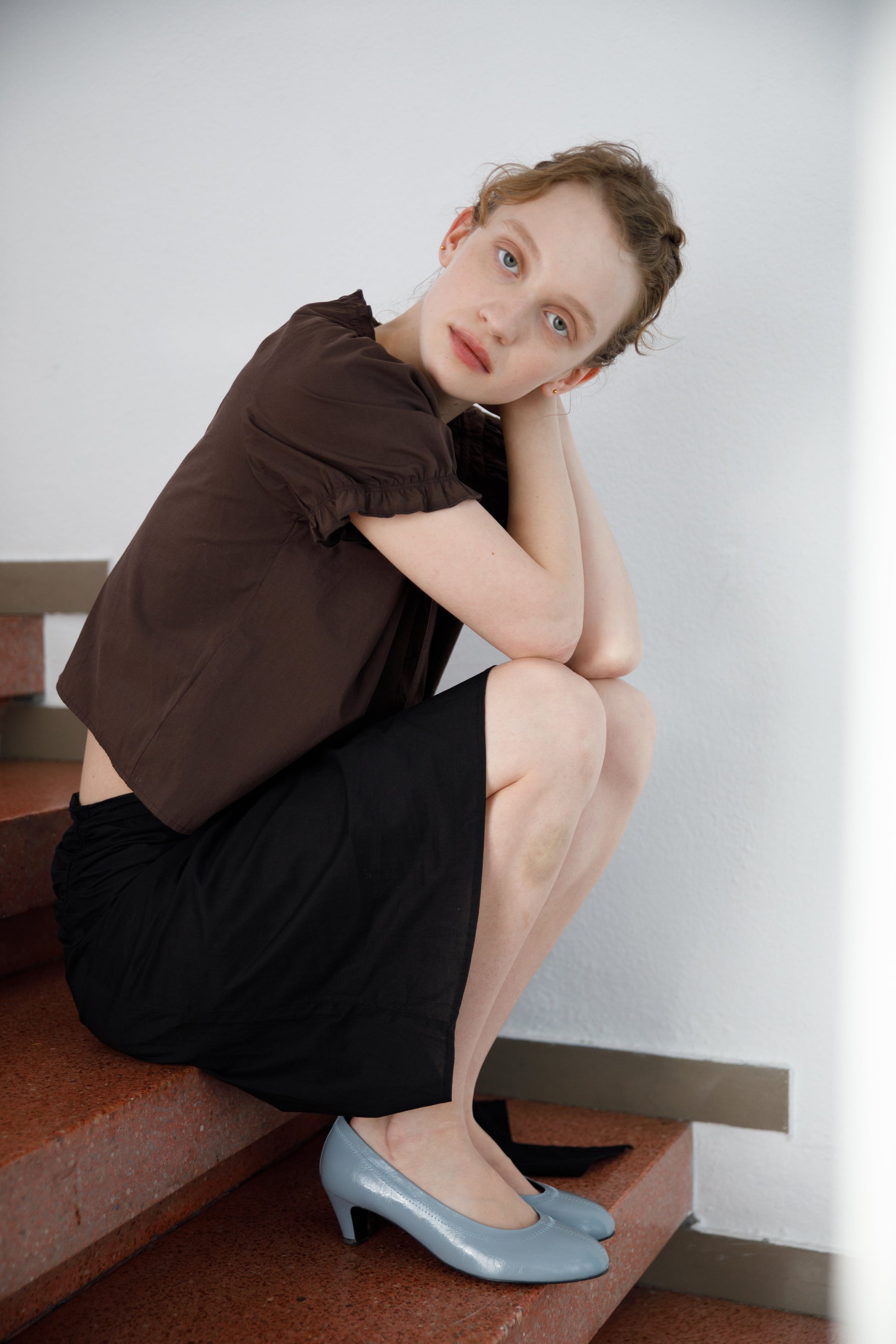A woman with light skin and blonde hair sits on red stairs, resting her chin on her knees. She wears the Close Shirt - Sepia by Deiji Studios, a black skirt, and light blue heels, gazing thoughtfully at the camera against a white wall.