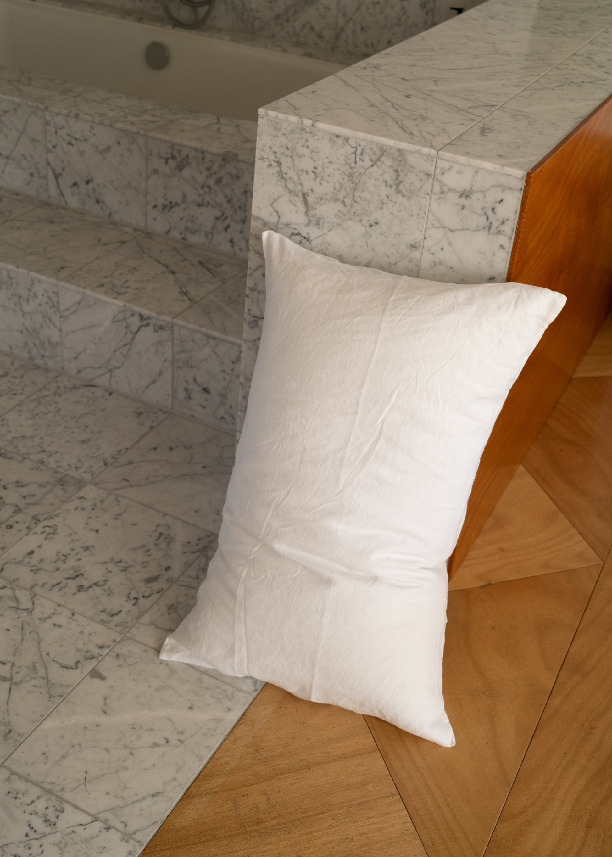 A white Pillow Slips cushion rests against marble steps and a wooden panel on a floor featuring a geometric wood design.