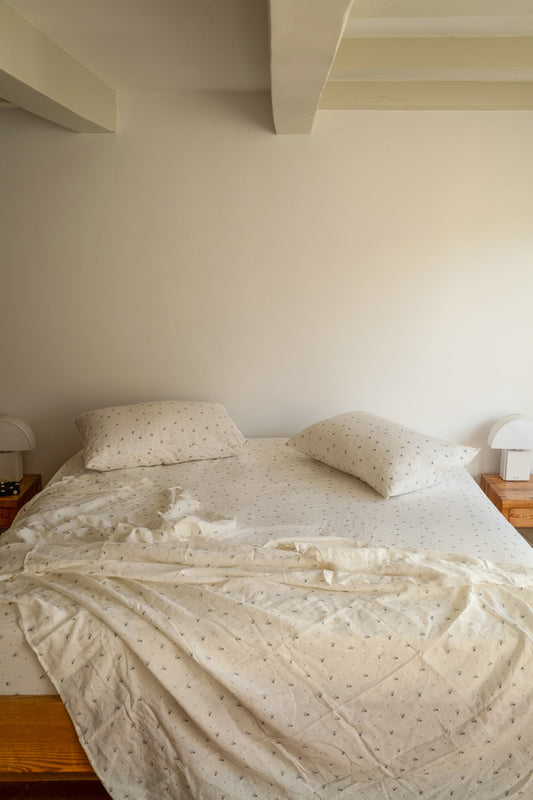 A bed with two pillows and patterned white Sheets sits against a cream wall, flanked by wooden nightstands with small white lamps on each side.