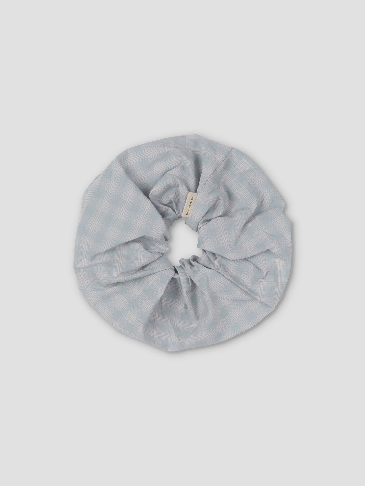 The Scrunchie is a large, light blue hair accessory with a subtle checkered pattern, shown on a plain light gray background.