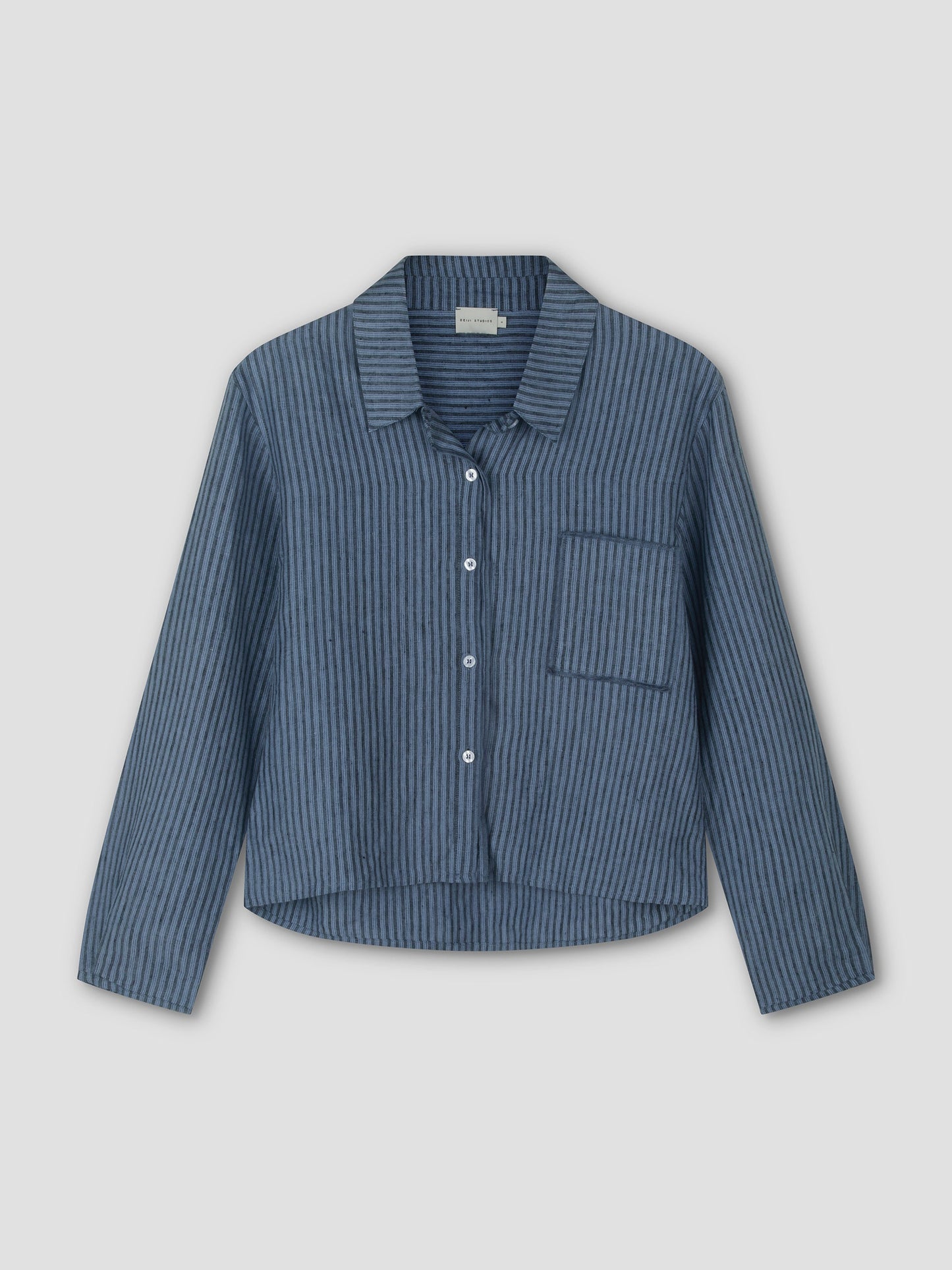 The Double Placket Shirt - Azure Stripe is a relaxed, long sleeve button-up with thin vertical stripes and a single chest pocket, shown against a plain light gray background.