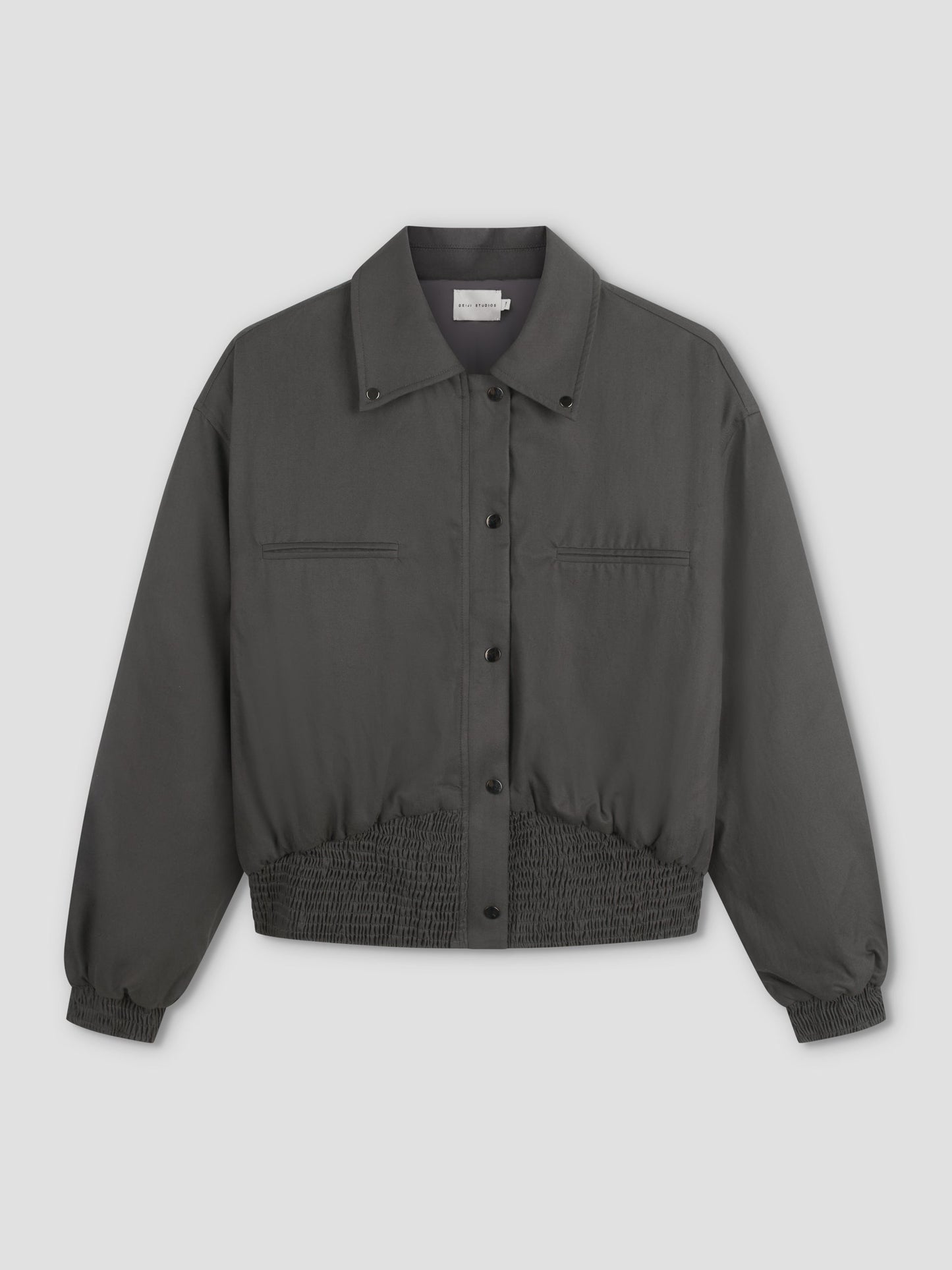 A Deiji Studios Shirring Jacket - Slate in dark gray with long sleeves, snap buttons, two chest pockets, and elasticated waistband and cuffs is displayed on a plain light background.