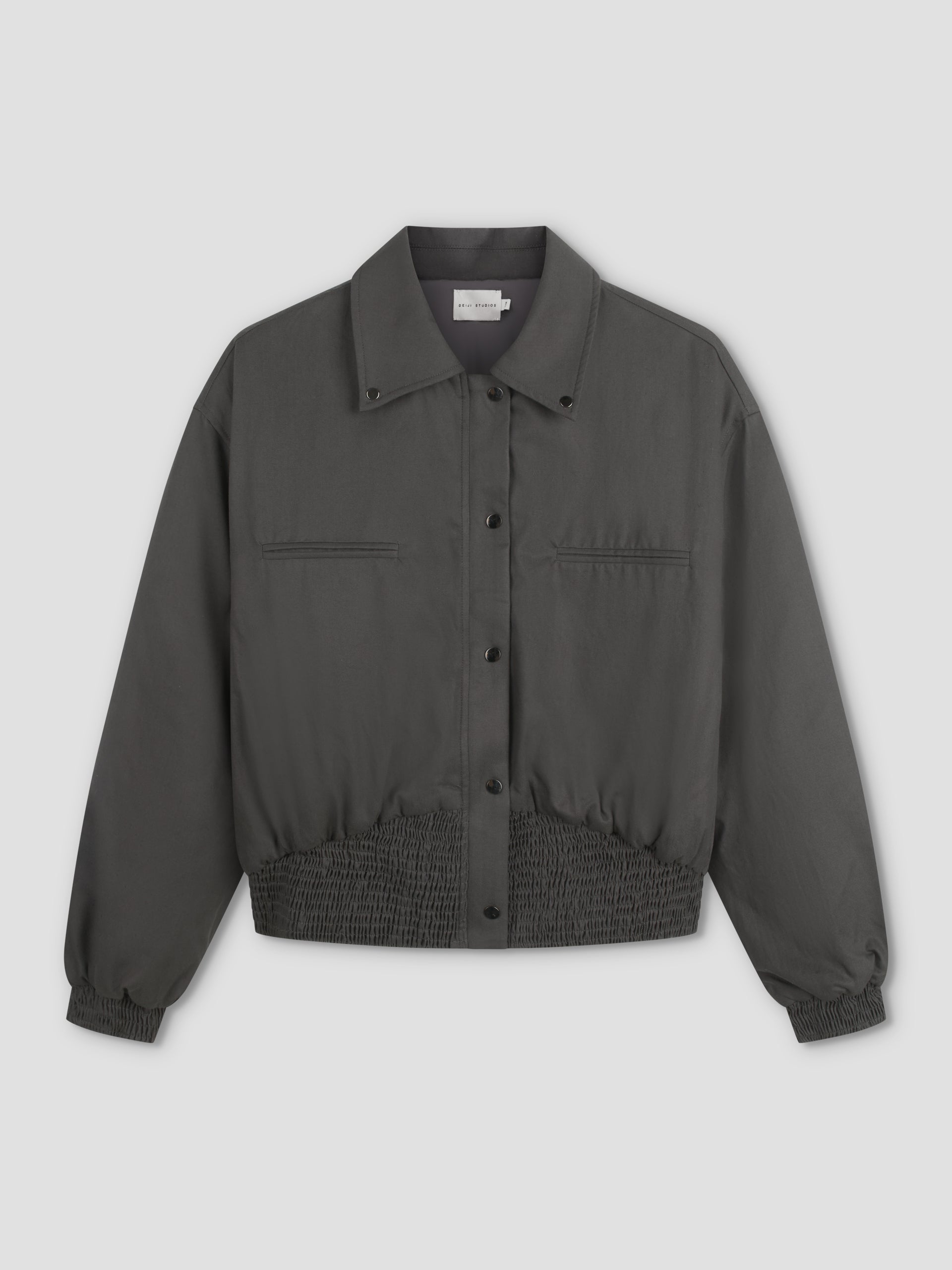 A Deiji Studios Shirring Jacket - Slate in dark gray with long sleeves, snap buttons, two chest pockets, and elasticated waistband and cuffs is displayed on a plain light background.