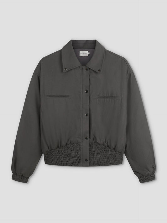 A Deiji Studios Shirring Jacket - Slate in dark gray with long sleeves, snap buttons, two chest pockets, and elasticated waistband and cuffs is displayed on a plain light background.