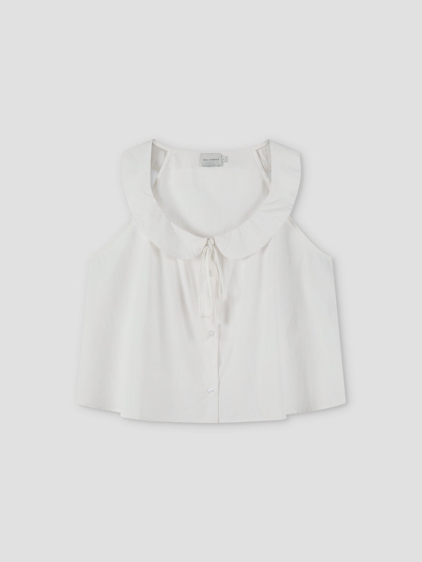 The Round Collar Top - White Texture by Deiji Studios is a sleeveless white top with a wide ruffled collar, front buttons, and a delicate bow tie at the neckline, shown against a plain light background.