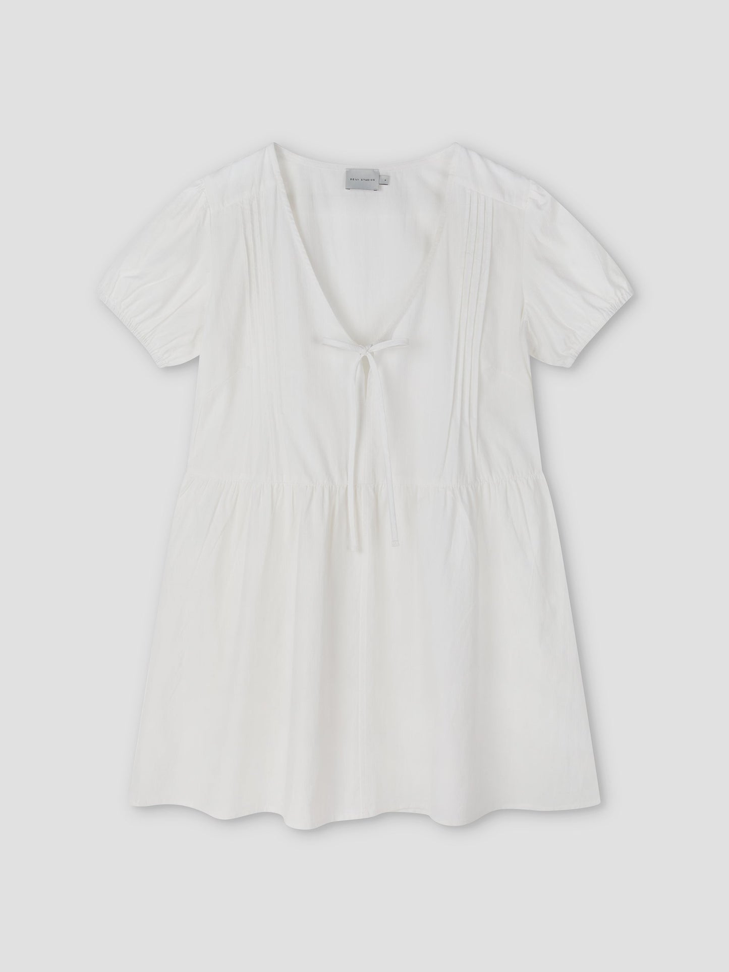 The V Pleat Dress - White Texture by Deiji Studios is a short-sleeve, lightweight white mini dress with a deep V-neckline, gathered waist, front tie detail, and subtle bodice pleats, shown laid flat on a light background.