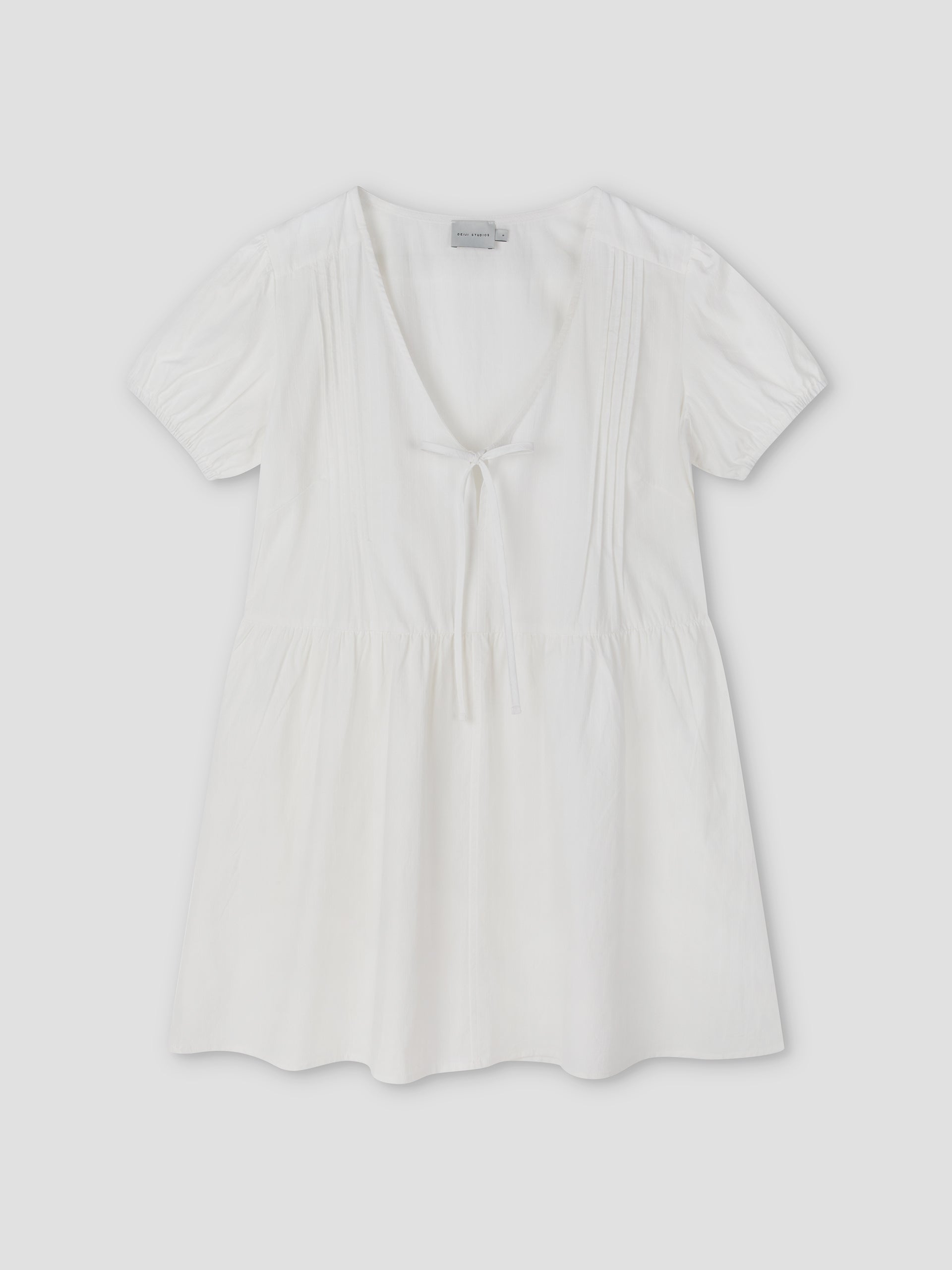 The V Pleat Dress - White Texture by Deiji Studios is a short-sleeve, lightweight white mini dress with a deep V-neckline, gathered waist, front tie detail, and subtle bodice pleats, shown laid flat on a light background.