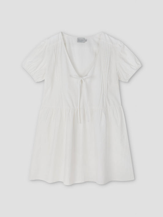 The V Pleat Dress - White Texture by Deiji Studios is a short-sleeve, lightweight white mini dress with a deep V-neckline, gathered waist, front tie detail, and subtle bodice pleats, shown laid flat on a light background.