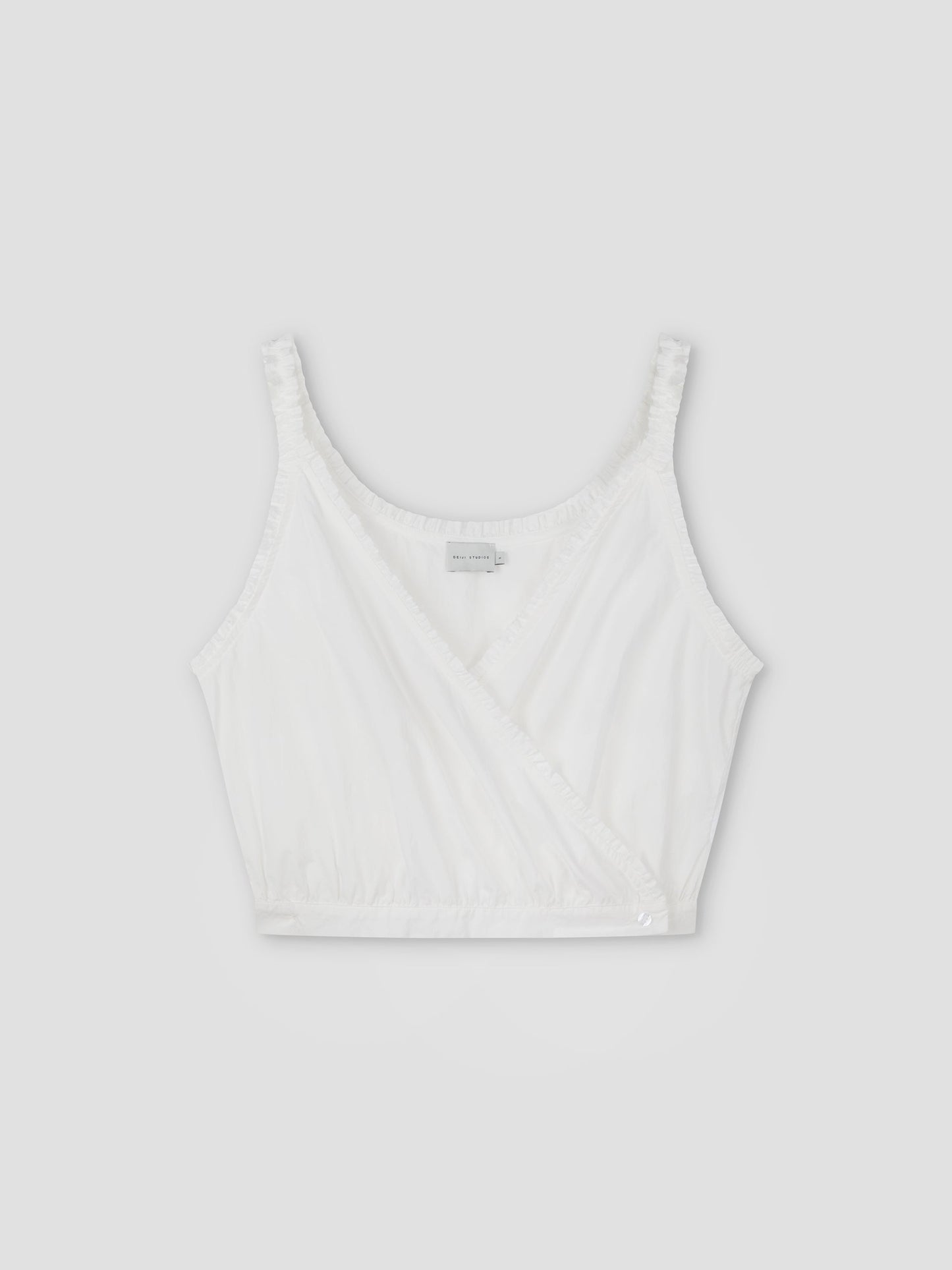 The Overlie Top - White features thin shoulder straps, a sleeveless wrap-style front, and gathered elastic at the hem, shown against a plain light gray background.