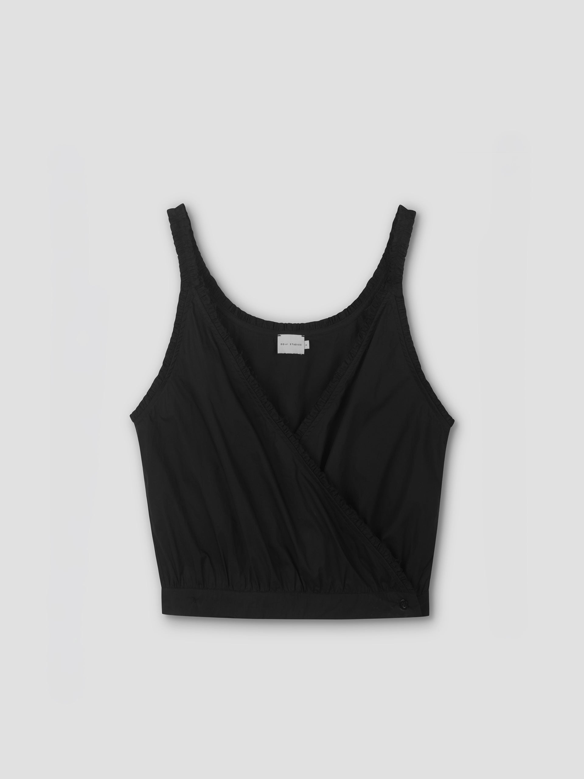 The Overlie Top - Black by Deiji Studios features a sleeveless, wrap-style front with a gathered hem and an interior label, shown against a light gray background.