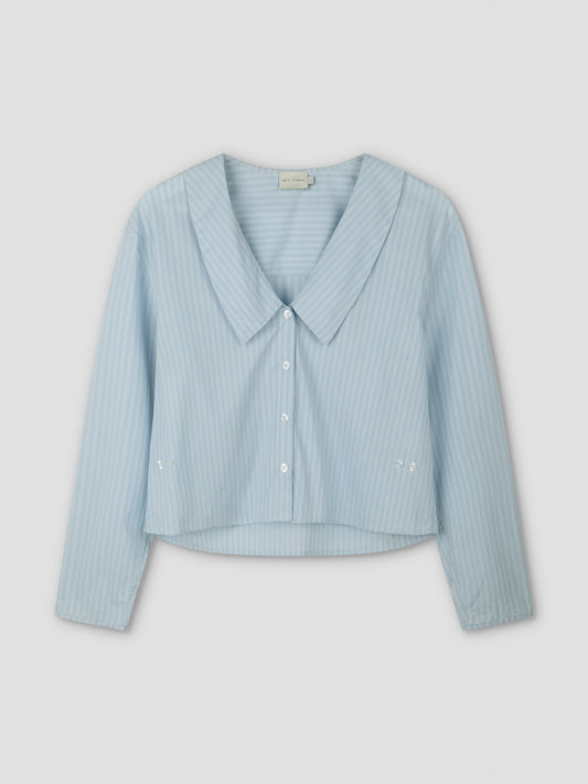 The Waist Button Shirt - Lake Blue Stripe features subtle vertical white stripes, a wide collar, cropped silhouette, long sleeves, and button-up front, displayed on a plain light background.