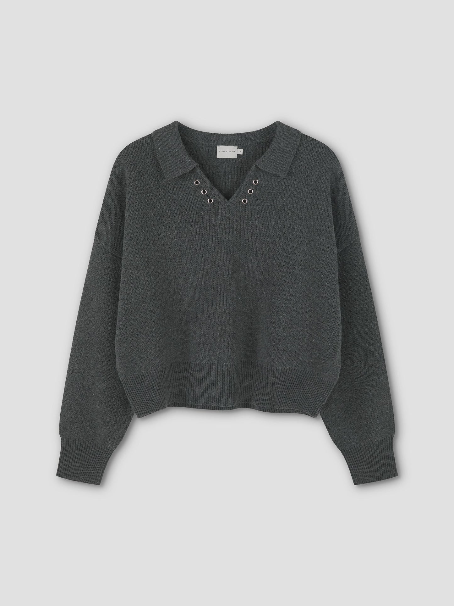 The Eyelet Knit - Dark Pebble by Deiji Studios is a dark gray relaxed long-sleeve sweater with a collar, V-neckline, and metal eyelets around the neck, shown laid flat on a plain light background.