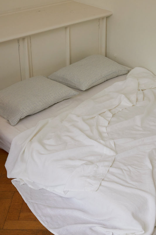 A bed showcases the Sheets - White with two gray-striped pillows, the breathable white sheet slightly rumpled, set against a white wall and wooden floor.