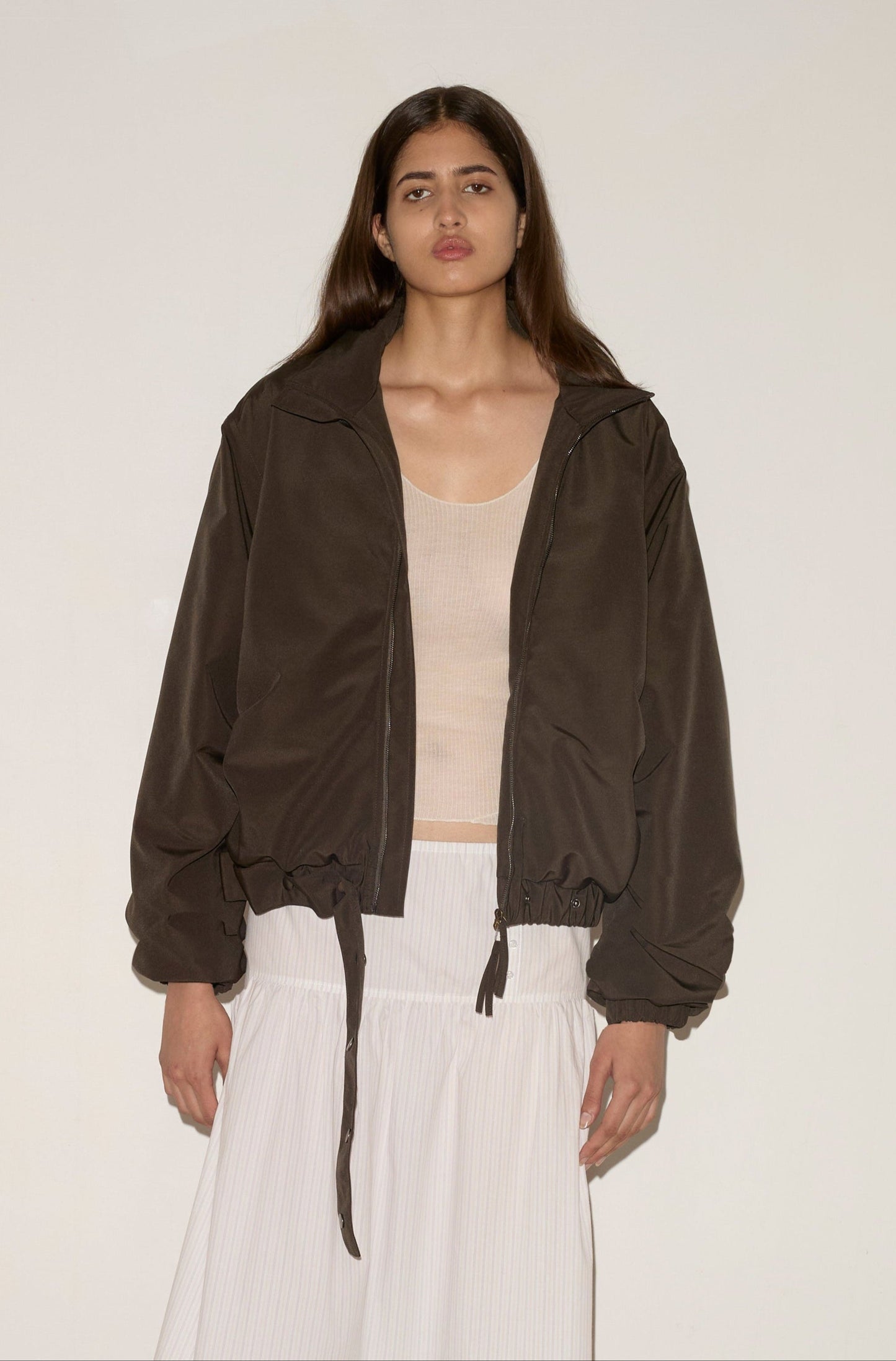 A woman with long brown hair models the 0 Press Stud Jacket in Dark Chocolate over a beige top and white pleated skirt, standing against a plain light-colored wall.