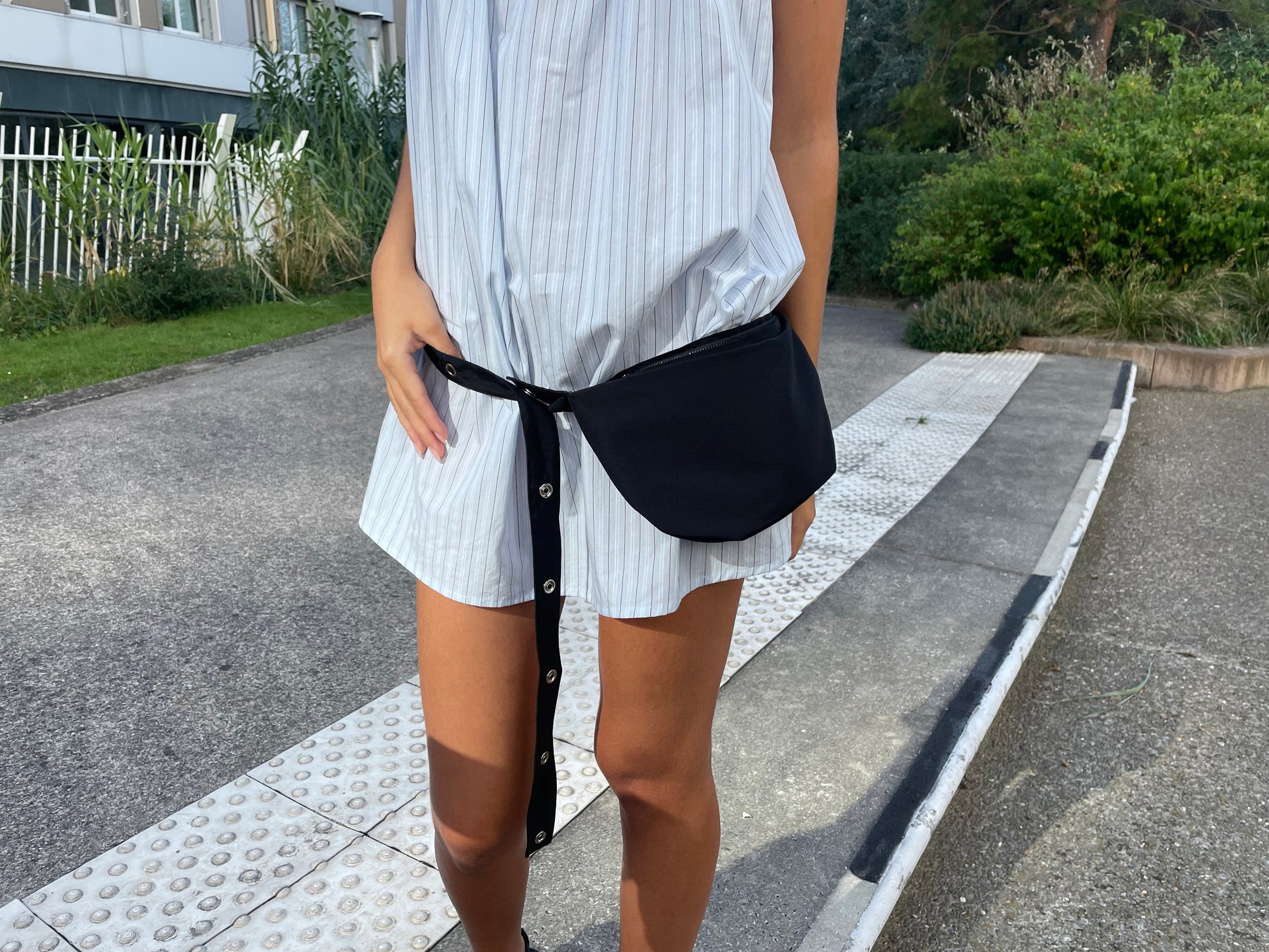 A person stands on a sidewalk wearing a light blue Deiji Studios striped shirt-dress and the Snap Belt Bag - Black, with one hand resting on the bag. Greenery and a building appear in the background.