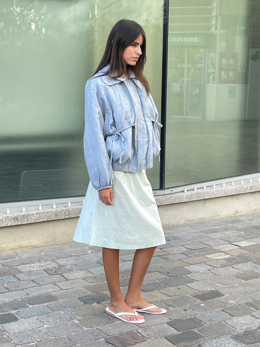 A woman with straight brown hair stands on a stone-paved sidewalk wearing a light blue jacket, the Simple Skirt - Mint, and white flip-flops, looking slightly to her left.