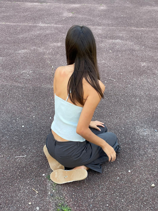 A woman with long brown hair, wearing the Fine Knot Tank in Ice Blue by Deiji Studios and dark pants, sits outdoors on textured brown ground with her back to the camera.