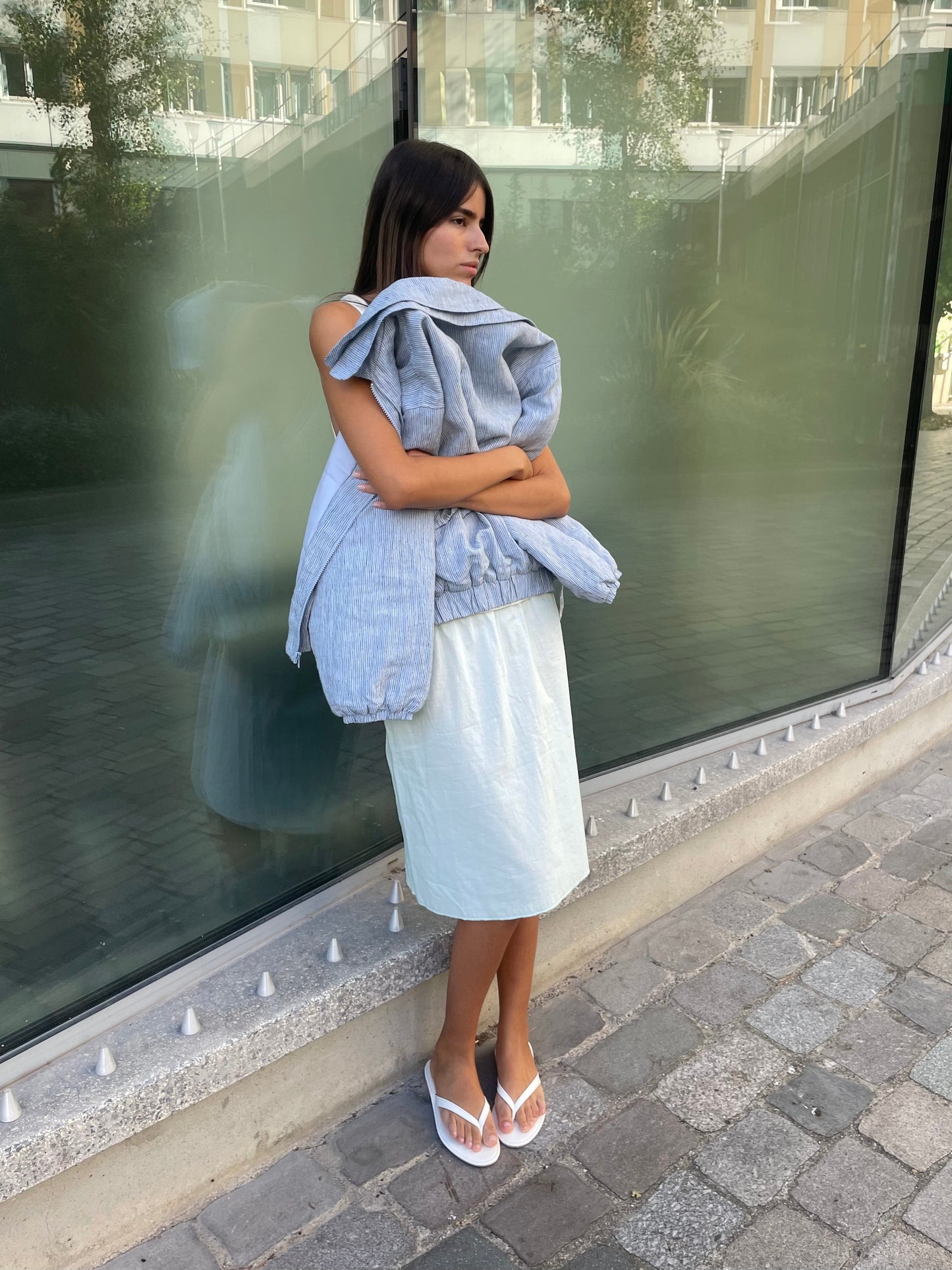 A woman stands outdoors by a glass window, wearing the Simple Skirt - Mint from Deiji Studios, white flip-flops, and holding a gray jacket. She looks to the side with arms crossed on a paved walkway.