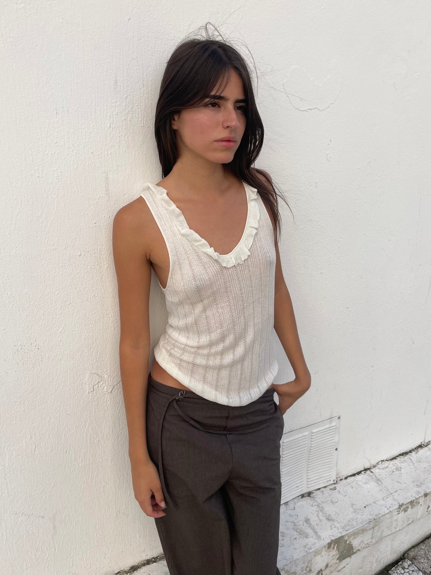 A woman with long dark hair wears a sleeveless white top with a ruffled neckline and Double Belted Pant - Brown Stripe, standing against a white textured wall with her hands in her pockets, gazing slightly to the side.
