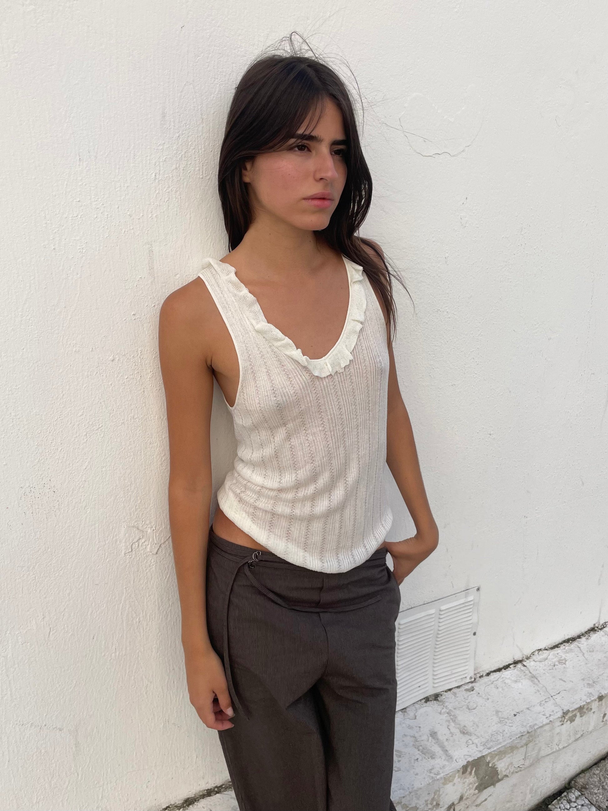 A woman with long dark hair wears a sleeveless white top with a ruffled neckline and Double Belted Pant - Brown Stripe, standing against a white textured wall with her hands in her pockets, gazing slightly to the side.