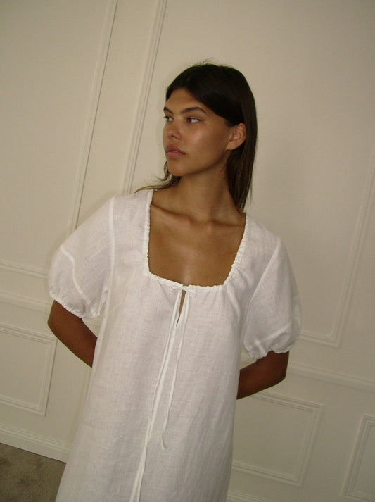 A woman with long brown hair stands against a cream-colored paneled wall, wearing the Curved Seam Midi Dress featuring short puff sleeves and a square neckline. She looks to the side with her hands behind her back.