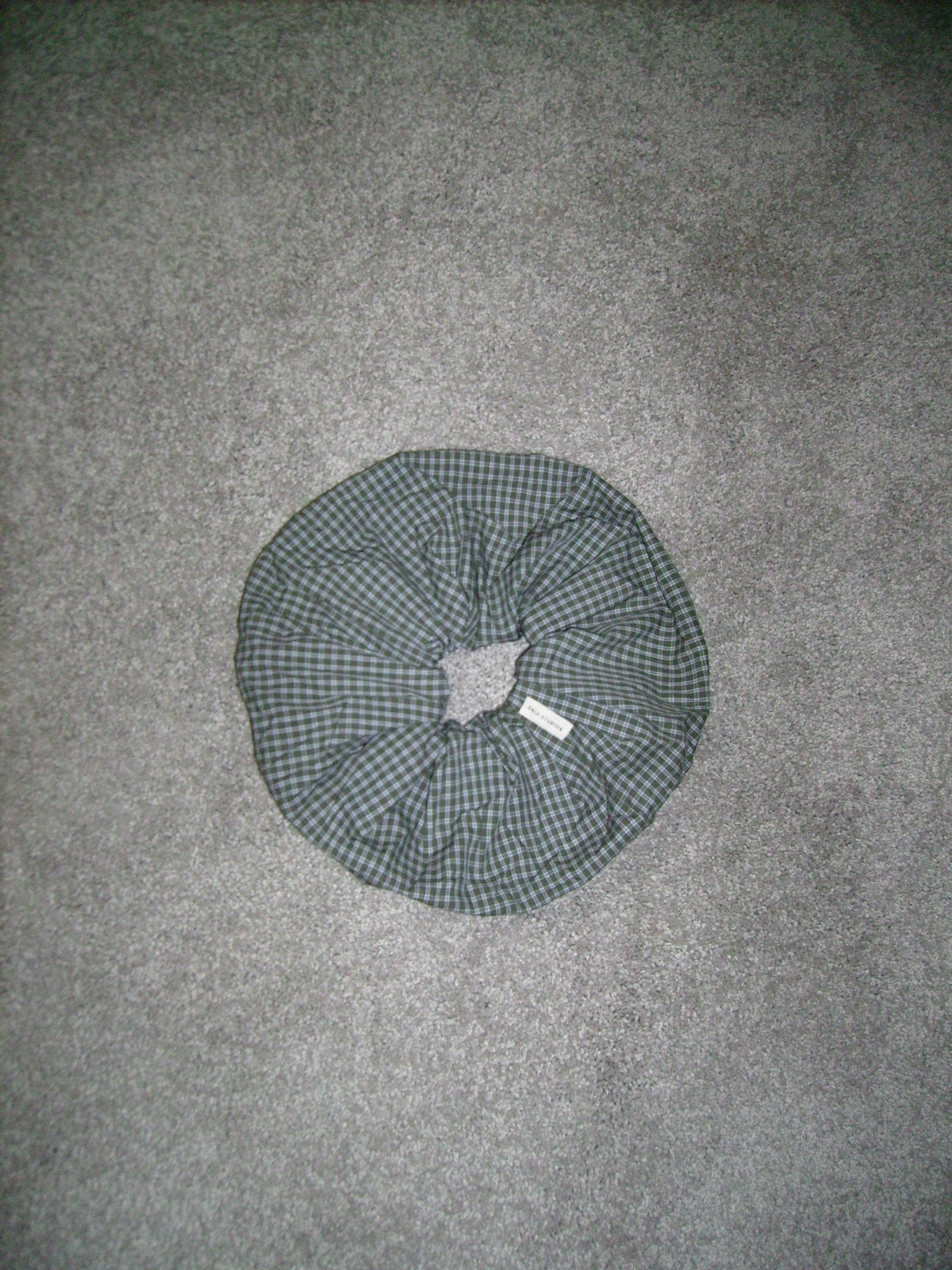 A round scrunchie with green and black plaid fabric and a center hole rests on a gray carpeted floor.