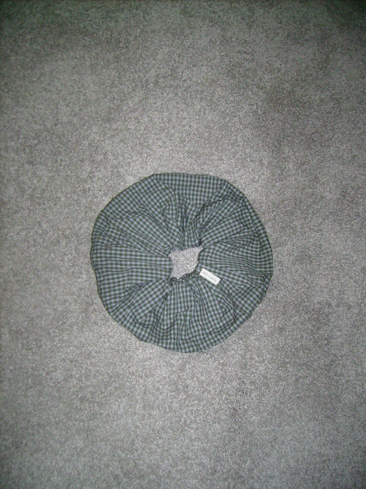 A round scrunchie with green and black plaid fabric and a center hole rests on a gray carpeted floor.