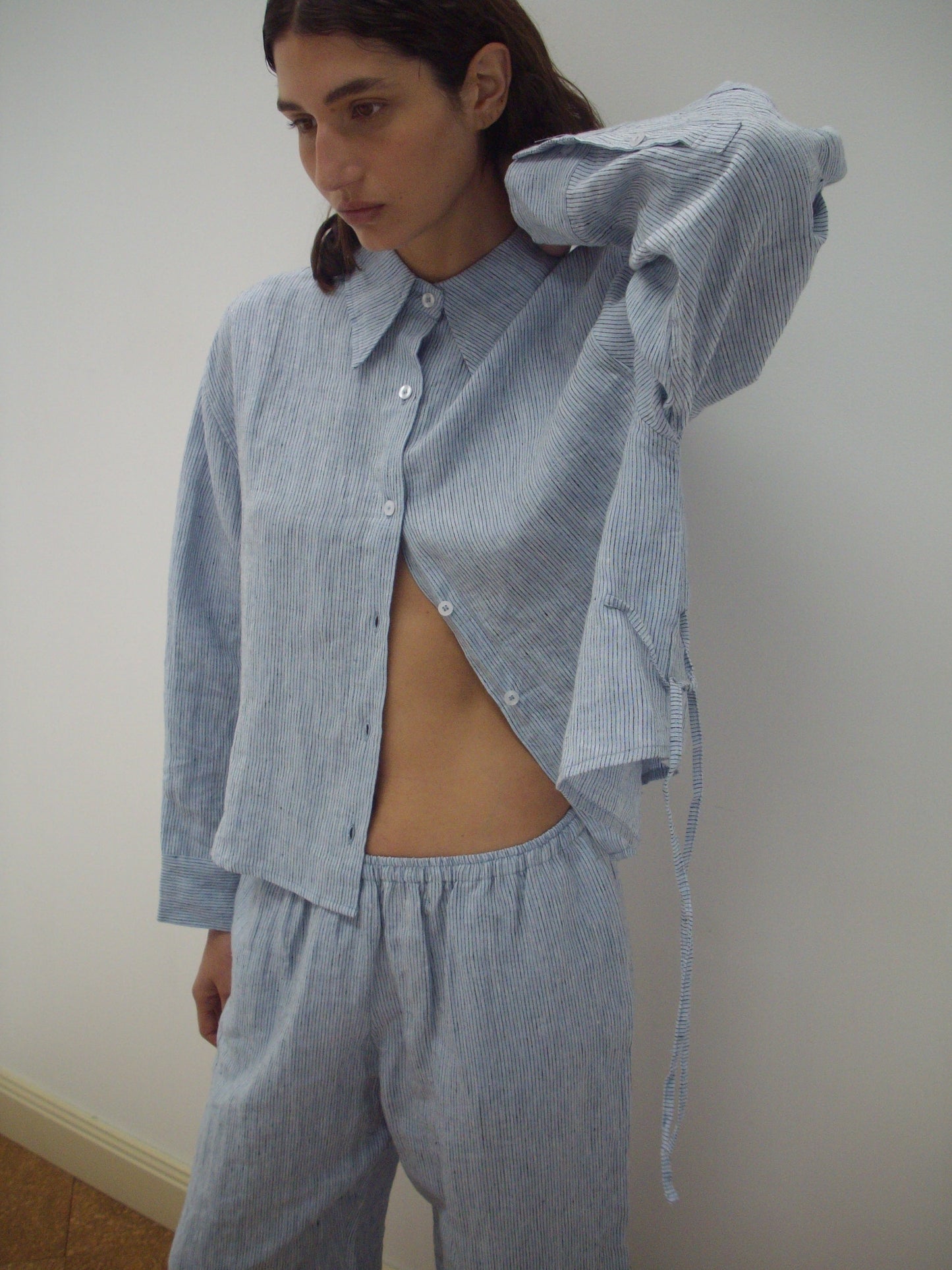 A person with long dark hair wears a loose, blue and white striped button-up shirt and matching pants from the 4 Tie Set. The shirt is partly unbuttoned as they stand against a plain wall, looking to the side.