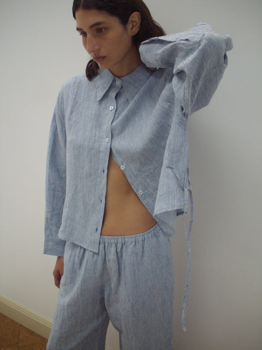 A person with long dark hair wears a loose, blue and white striped button-up shirt and matching pants from the 4 Tie Set. The shirt is partly unbuttoned as they stand against a plain wall, looking to the side.