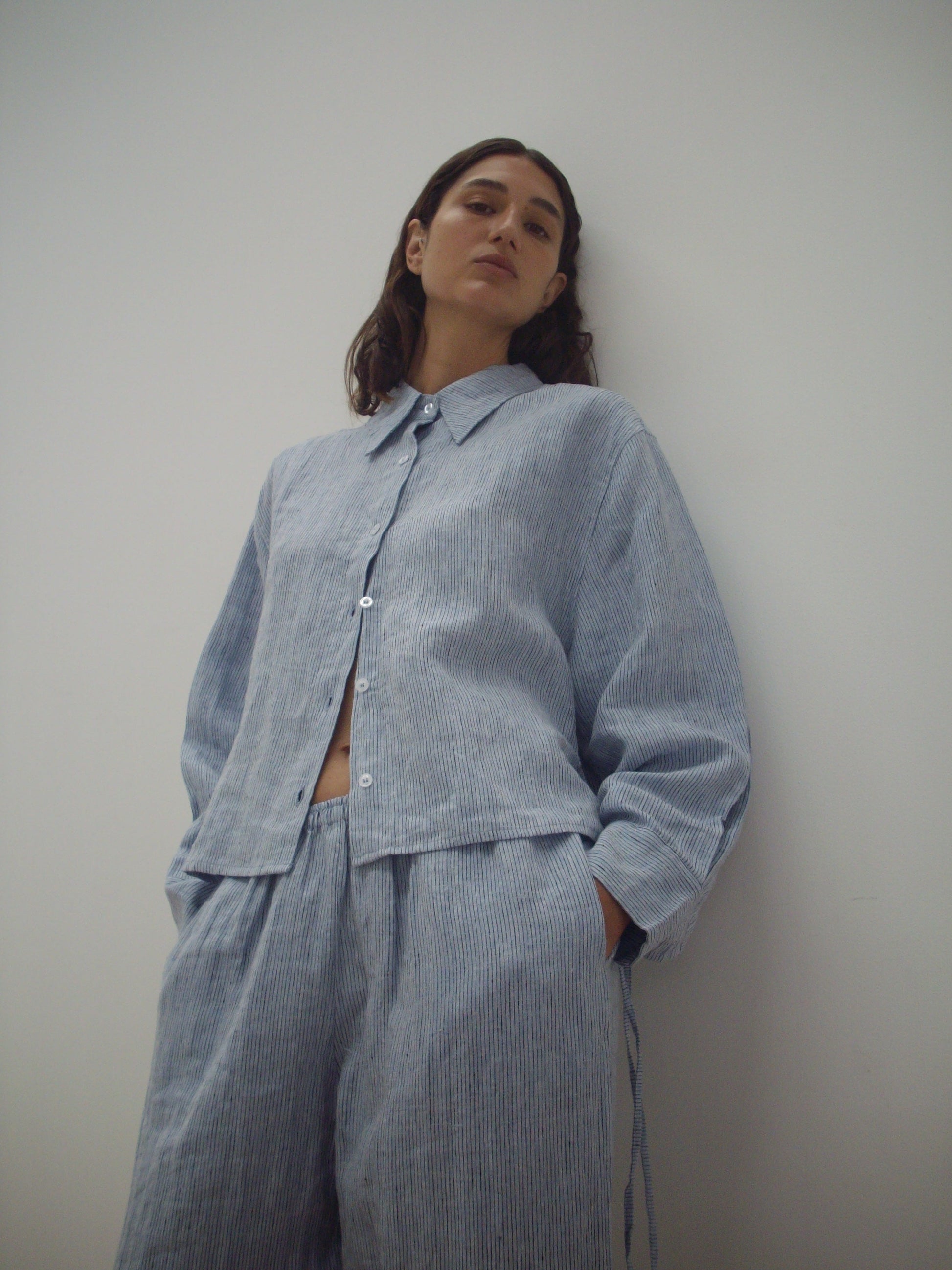 A person stands against a plain white wall, wearing Deiji Studios' 4 Tie Set - Blue Stripe: a loose-fitting, light blue and white striped organic shirt with matching wide-leg pants, looking confidently at the camera.