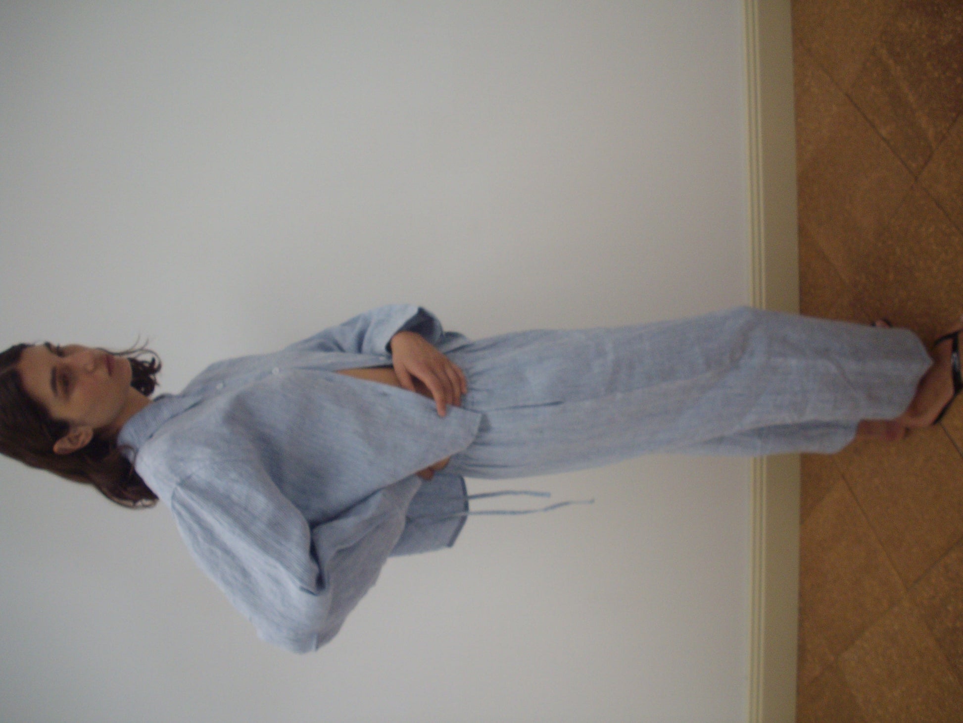 A person stands sideways against a plain white wall, wearing the 4 Tie Set - Blue Stripe by Deiji Studios, a loose-fitting French linen pantsuit with long sleeves and wide-leg pants, paired with open-toe sandals.