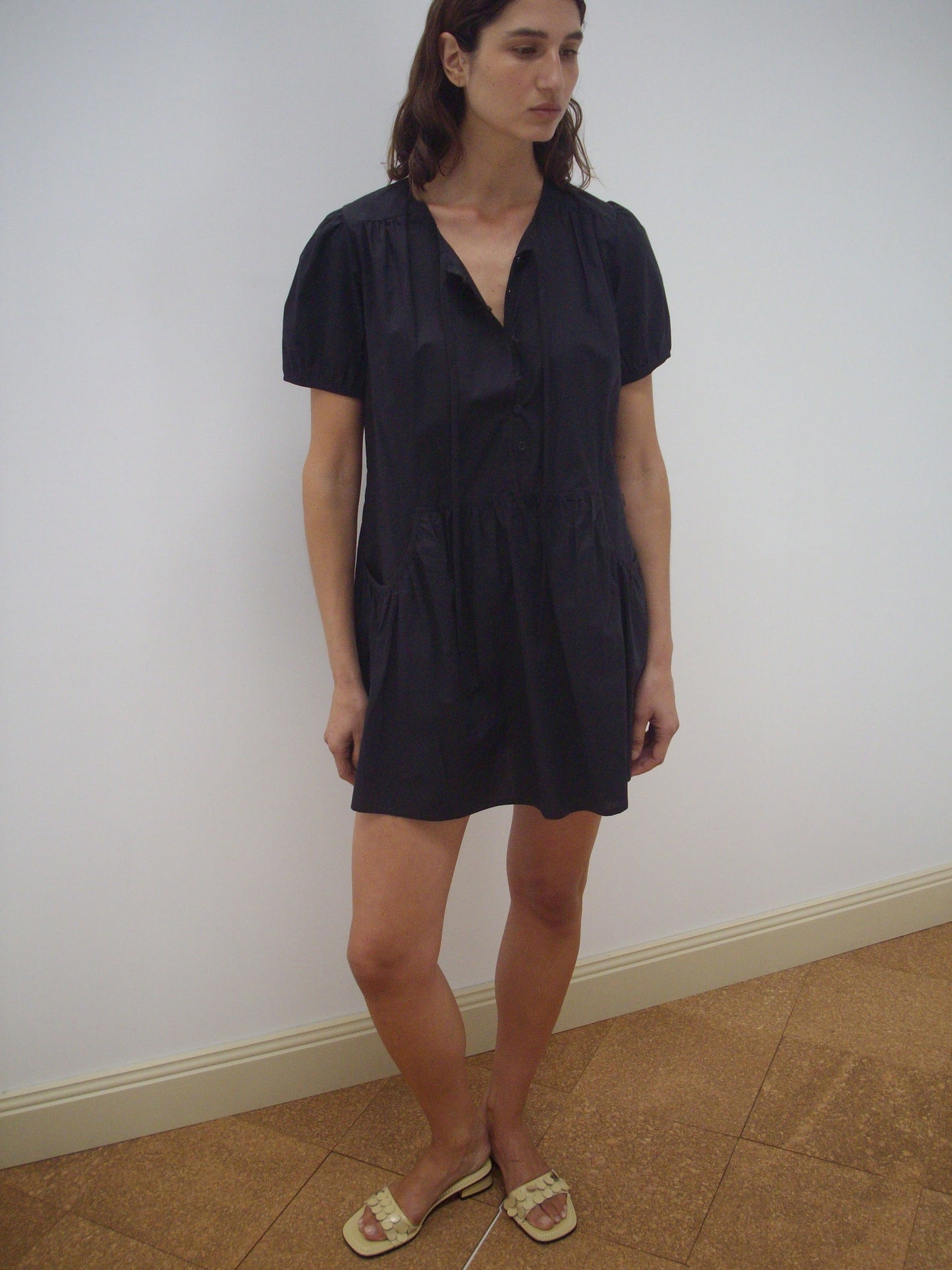 A woman stands indoors against a white wall, wearing the Pocket Mini Dress - Black by Deiji Studios and beige sandals. She has straight brown hair, a neutral expression, and one leg slightly bent.