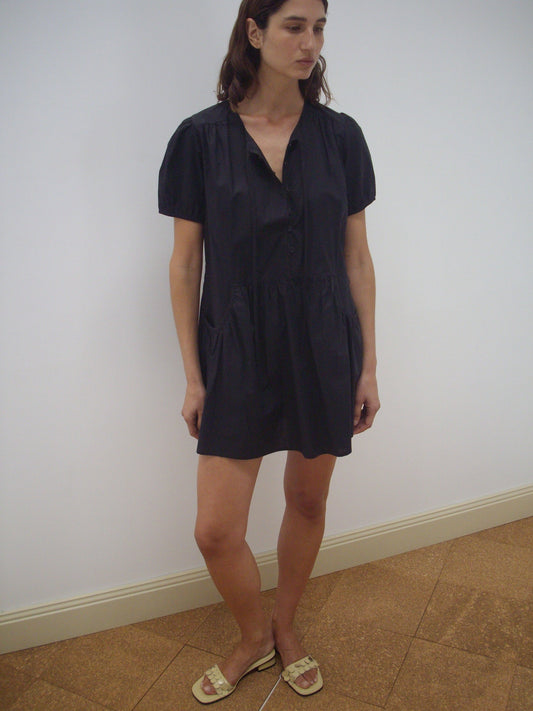 A woman stands indoors against a white wall, wearing the Pocket Mini Dress - Black by Deiji Studios and beige sandals. She has straight brown hair, a neutral expression, and one leg slightly bent.
