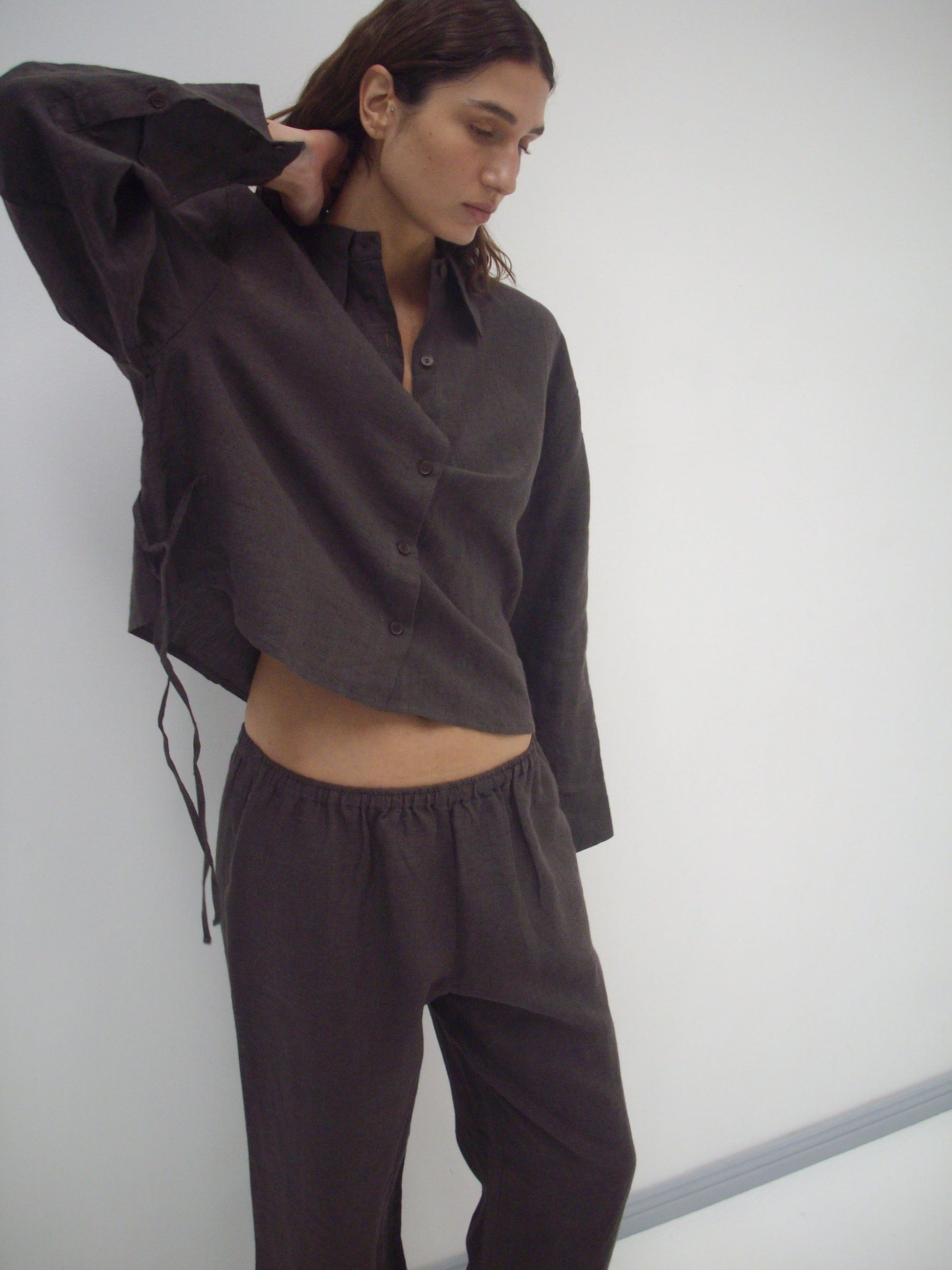 A person wears the Deiji Studios 4 Tie Set - Bark, a loose organic linen outfit in dark brown, standing against a plain white background with one arm raised and head slightly tilted down.