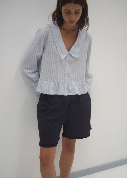 A woman stands against a white wall, wearing the Sweep Shirt—a light blue, long-sleeved, pinstriped blouse with a wide collar and ruffled hem—paired with loose black shorts. She looks down thoughtfully, hands in pockets.