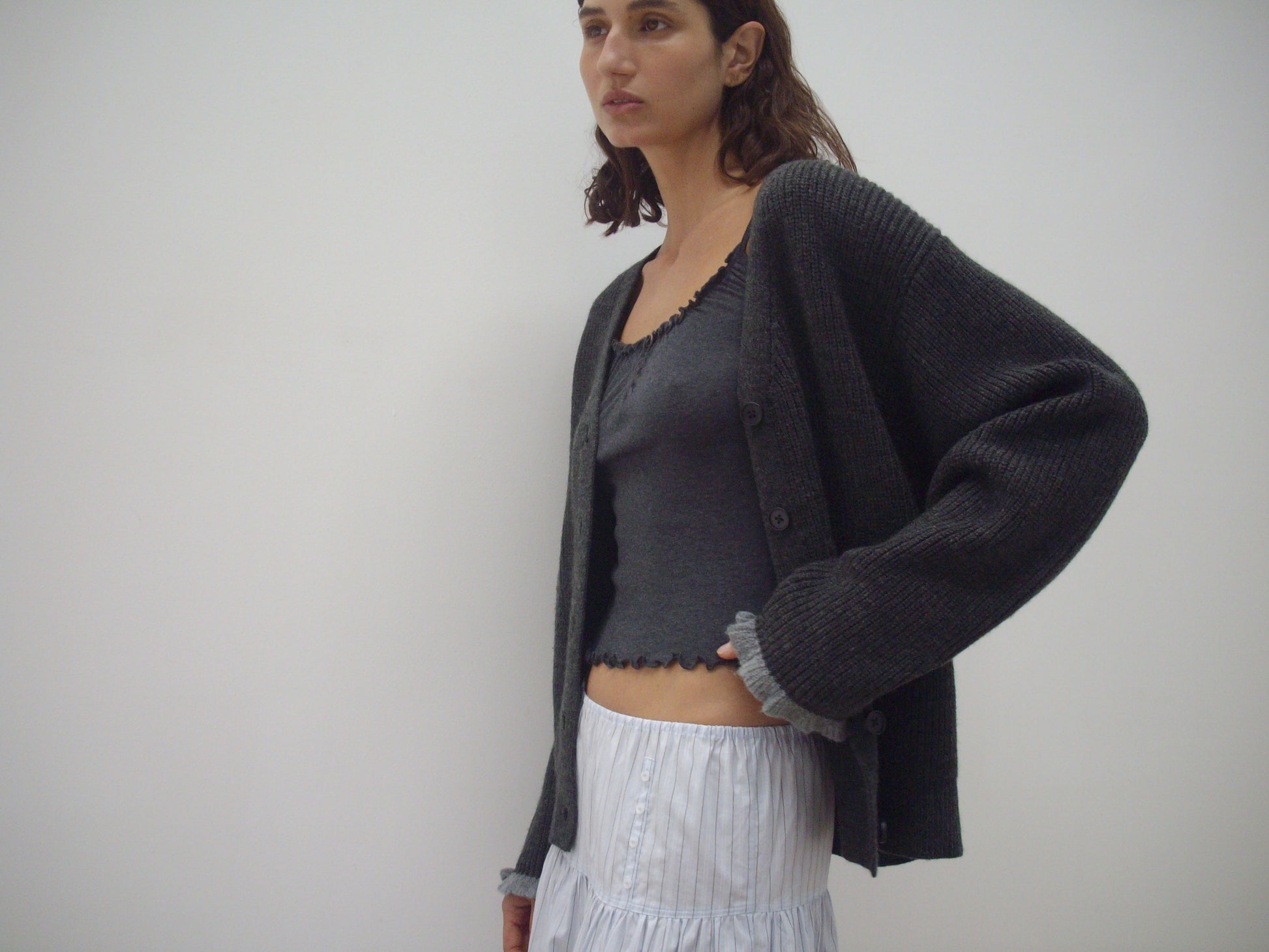 A woman stands before a plain light background, wearing the Flounce Cardigan - Charcoal by Deiji Studios over a matching ruffled tank top and a white tiered skirt.