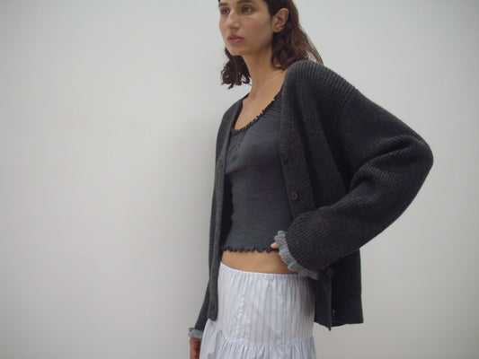 A woman stands before a plain light background, wearing the Flounce Cardigan - Charcoal by Deiji Studios over a matching ruffled tank top and a white tiered skirt.